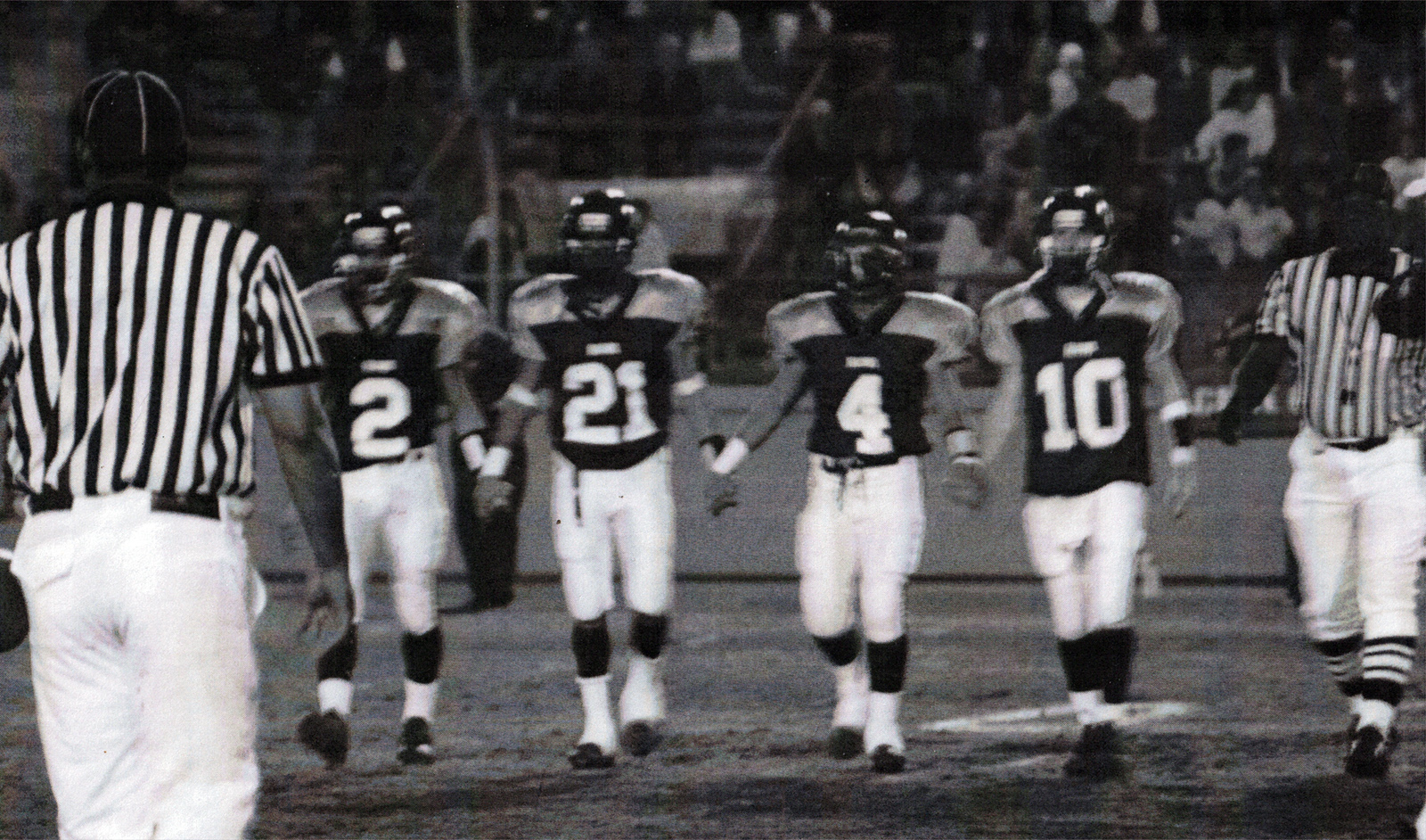 Black and white photo of four football players walking on the field.